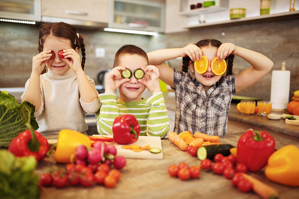 Trois enfants s'amusant avec des légumes et des fruits, utilisant des tranches de tomates, de concombres et d'oranges comme lunettes dans une cuisine colorée | Le Petit Décante
